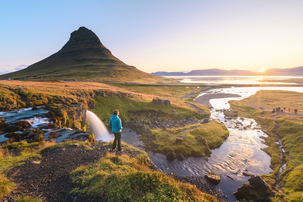 Mann står ved stor bekk mens han ser mot solnedgang. Spiss fjelltopp i bakgrunnen