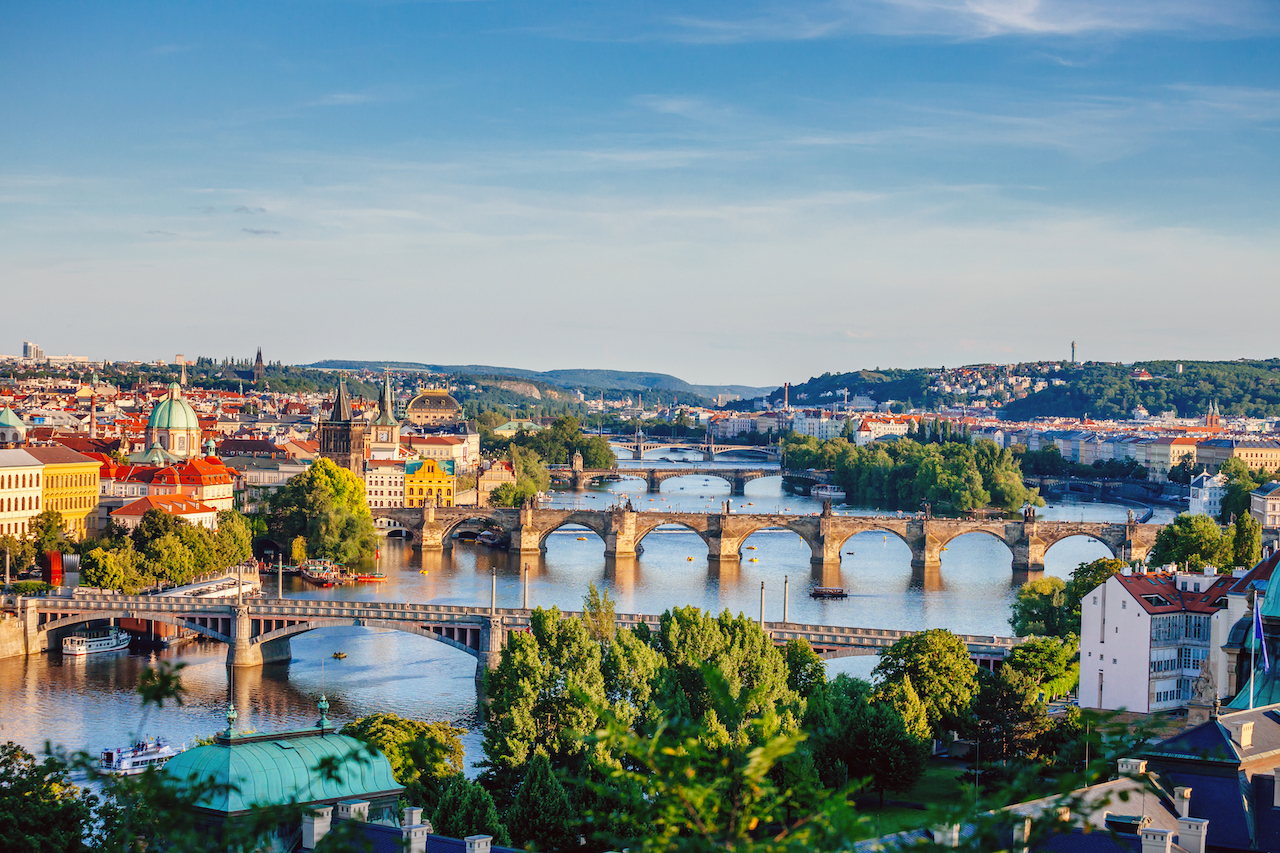 Elven Vltava omkranset av vakre bygg i solskinn, og flere broer som krysser elven.