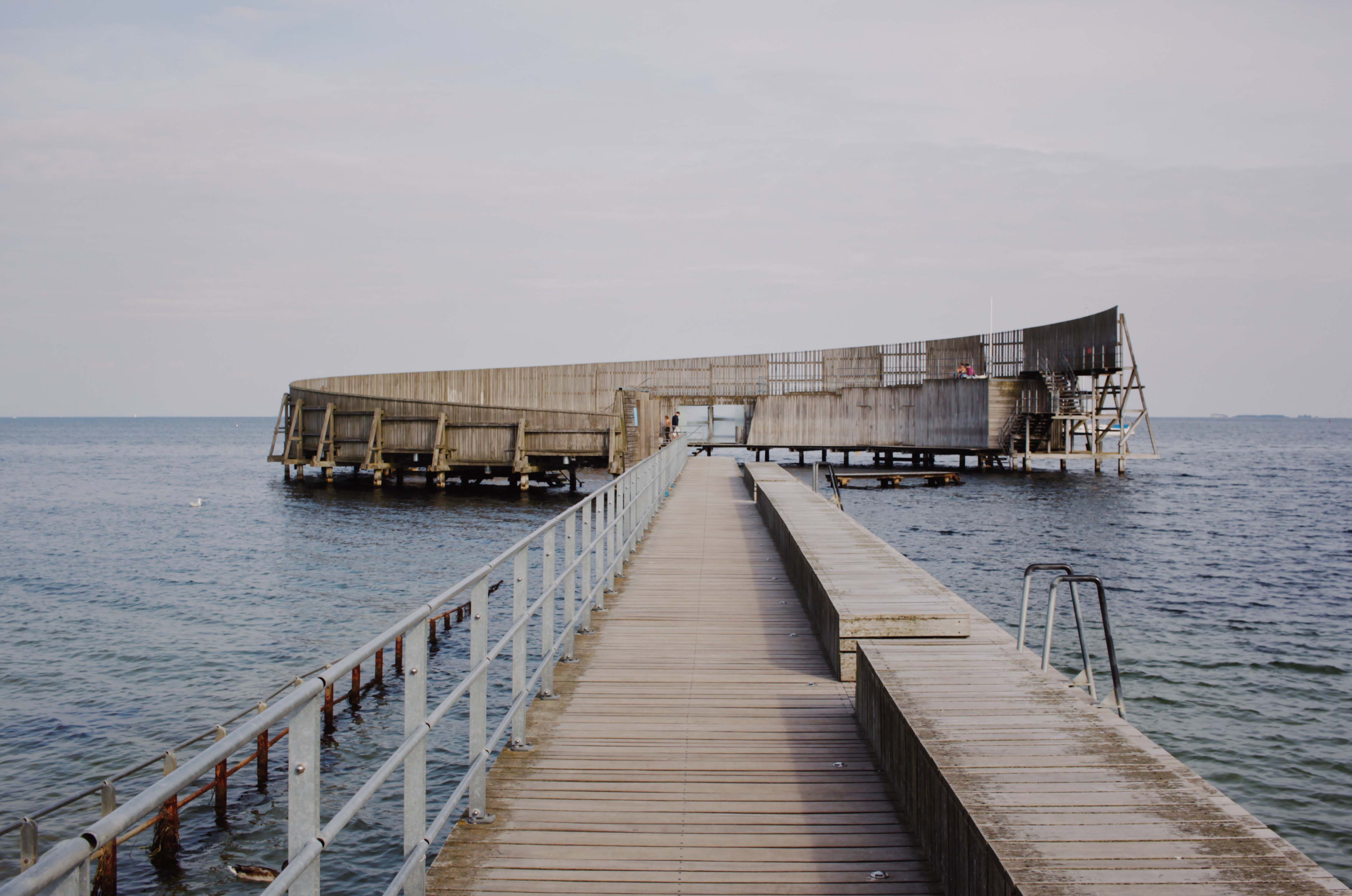 Brygge ut mot sjøen som leder ut til et rundt, modernistisk badehus i treverk