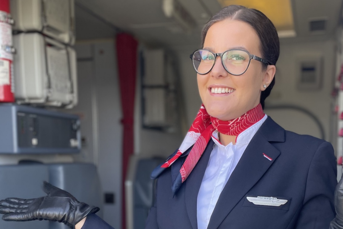 Female airline crew with brown hair in a ponytail and glasses, smiles as she stands in the entrance of airplane