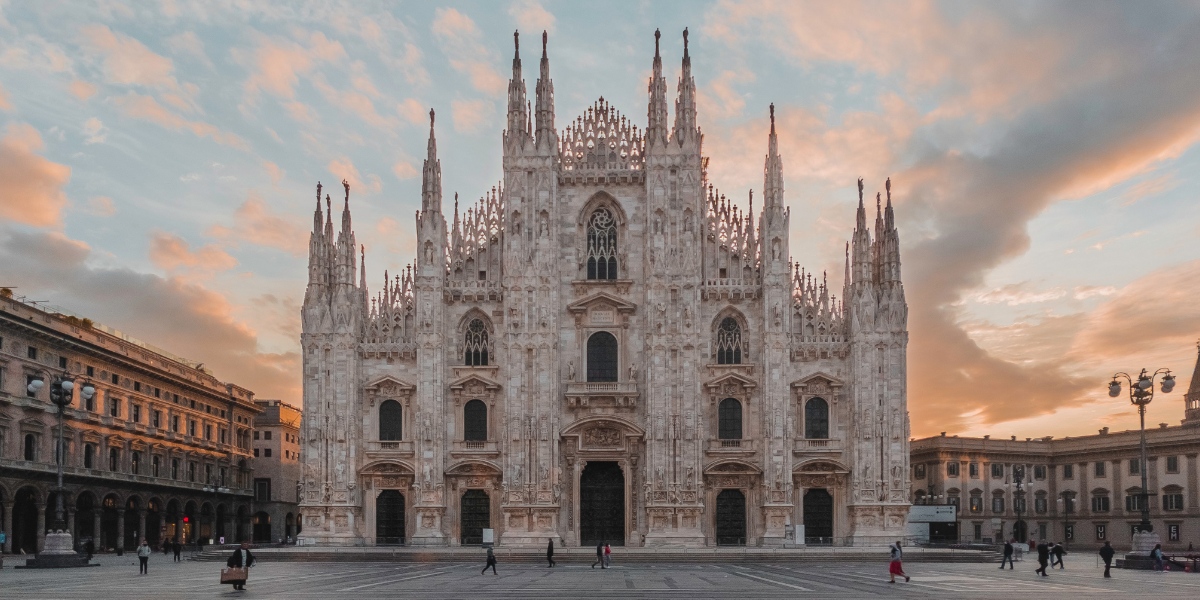 katedralen i milano med sine spir og karakteristiske form i hvit markmor. plassen foran er tom. Solen er i ferd med å gå ned bak kirken.