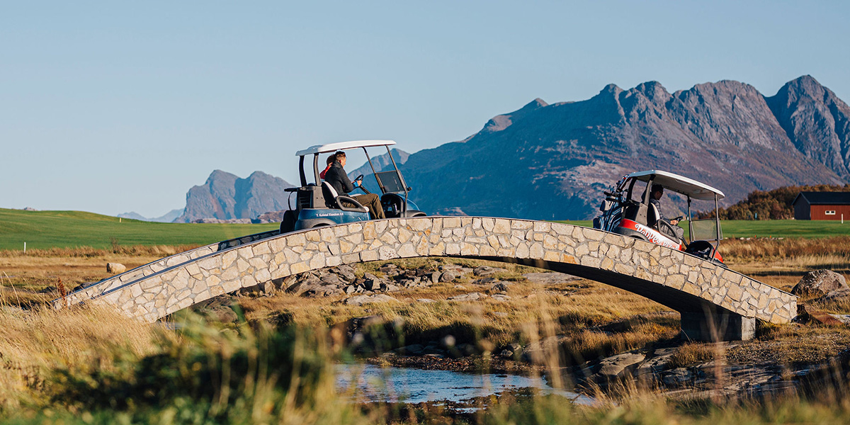To golfbiler kjører over en liten steinbro på en golfbane, med fjell i bakgrunnen