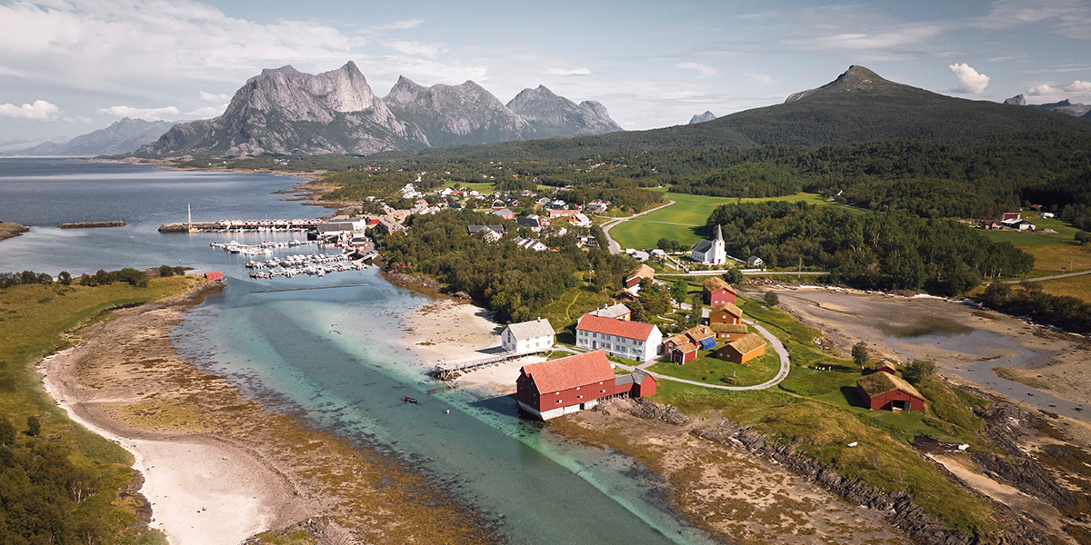 Gamle gårdsbygninger sett fra fugleperspektiv med fjell, sjø og strand rundt