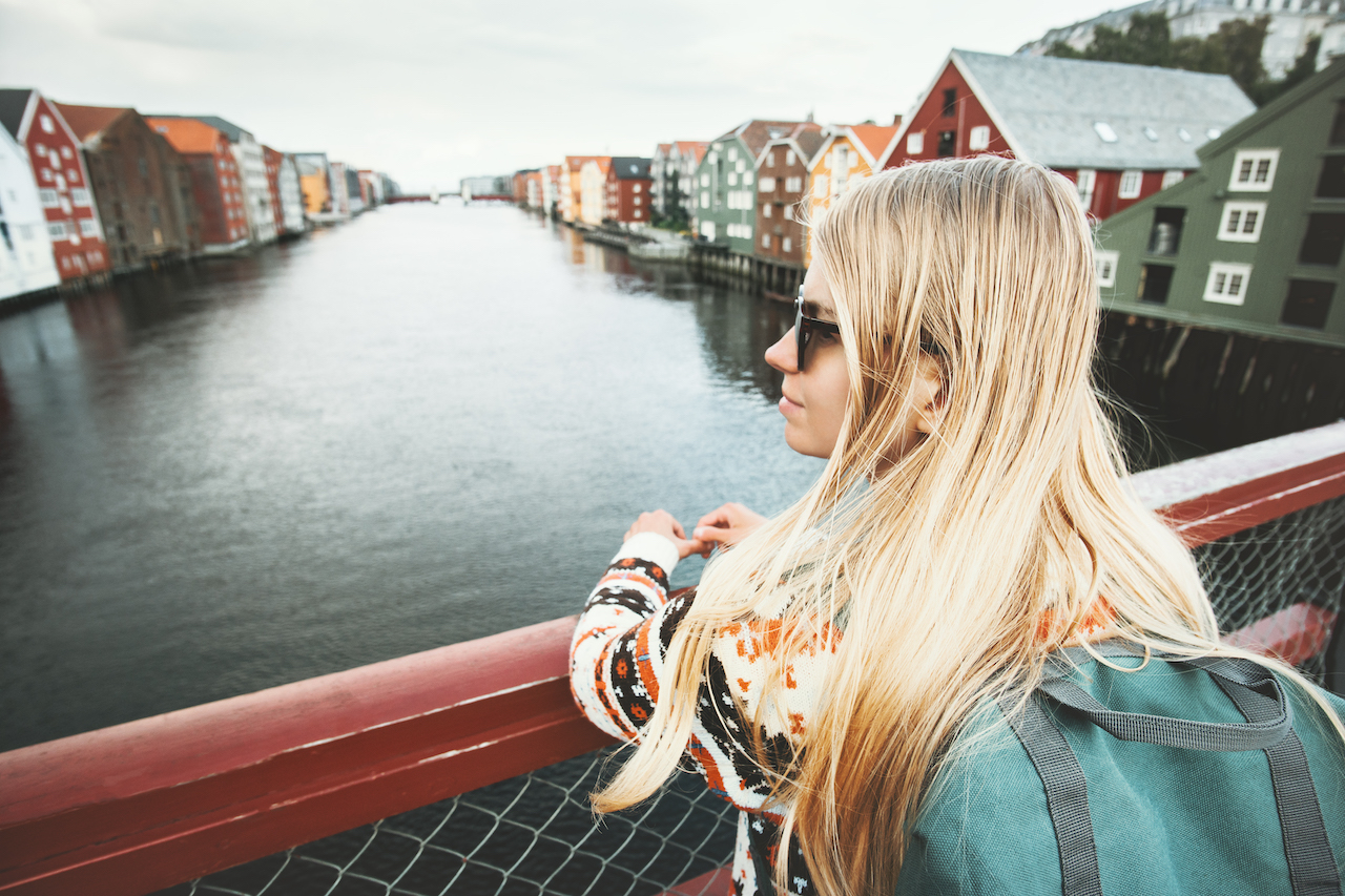 Ung kvinne med langt blondt hår og ryggsekk står på en bro og ser utover Nidelven